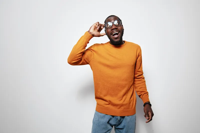 African American man with dark skin wearing orange sweater and glasses smiling and posing on white background. Casual style, happy expression and modern fashion for young adult male.