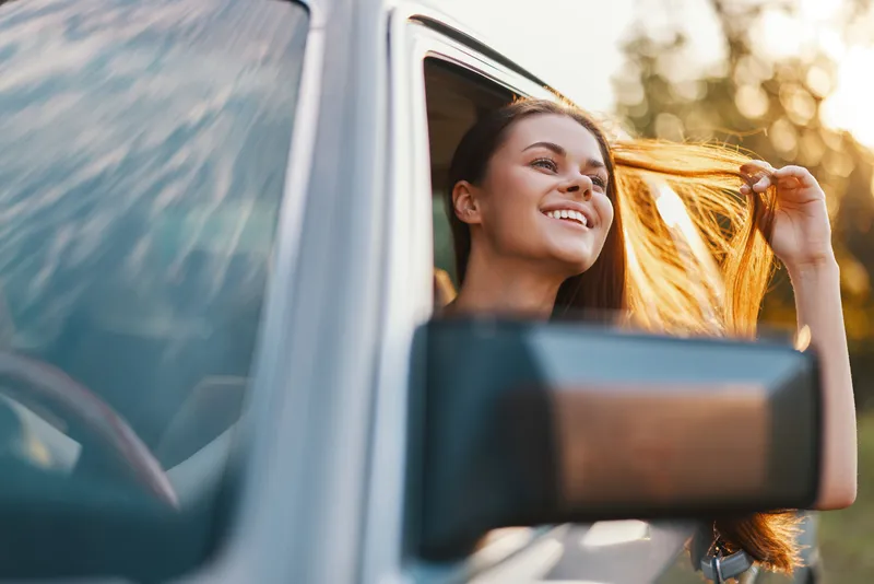 Joyful young woman enjoying a road trip with wind in her hair, carefree expression, natural backdrop and warm sunlight highlighting her happiness.