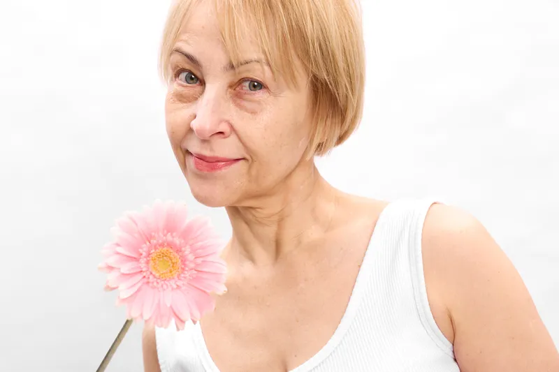 Blonde elderly woman holding pink gerbera flower, close up portrait, soft smile, white background, studio daylight, gentle beauty, calm mood, minimal composition, quiet atmosphere