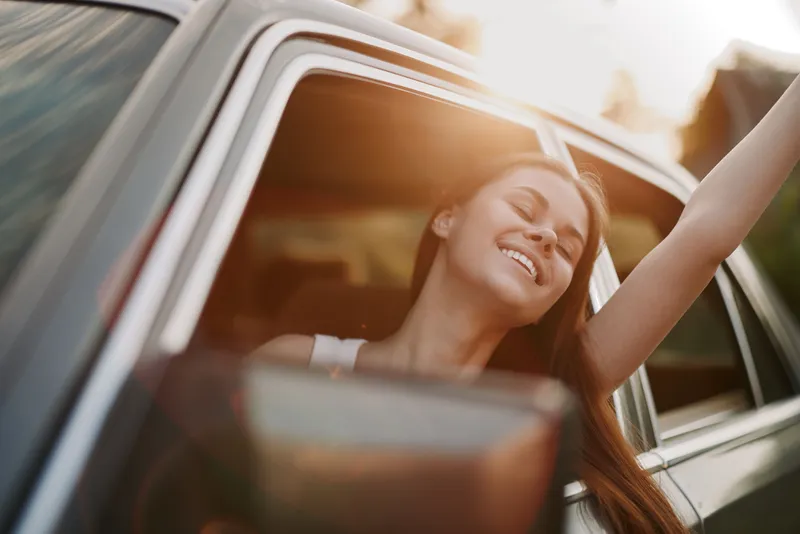 A joyful young woman enjoying a sunny day while leaning out of a car window, celebrating freedom and adventure with a bright smile.