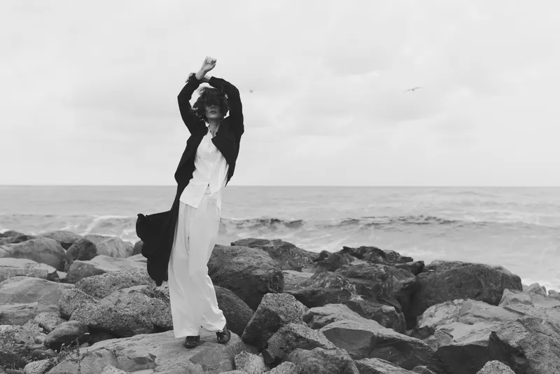 Dancer on a rocky shore by the sea wears a flowing white dress and dark cardigan, captured in monochrome, conveying movement, freedom, and a timeless coastal mood