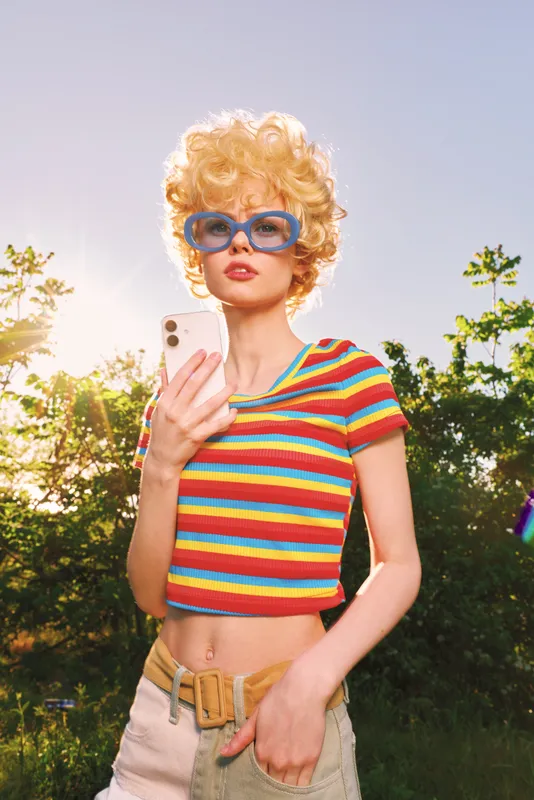 A young woman with curly blonde hair and blue sunglasses poses in a sunny park, wearing a colorful striped crop top and beige shorts, exuding playful confidence in the bright outdoors.