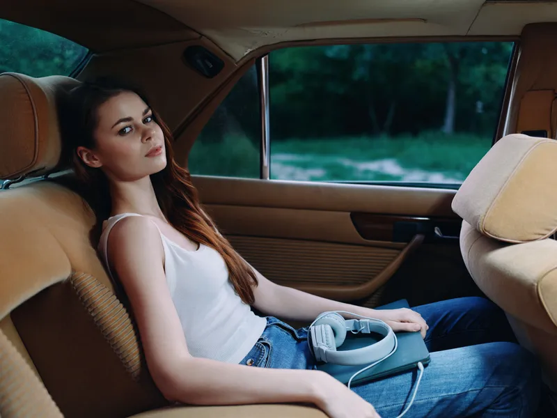 Young woman relaxing in a vintage car, wearing casual clothes, headphones on, immersed in music amidst a serene outdoor background.