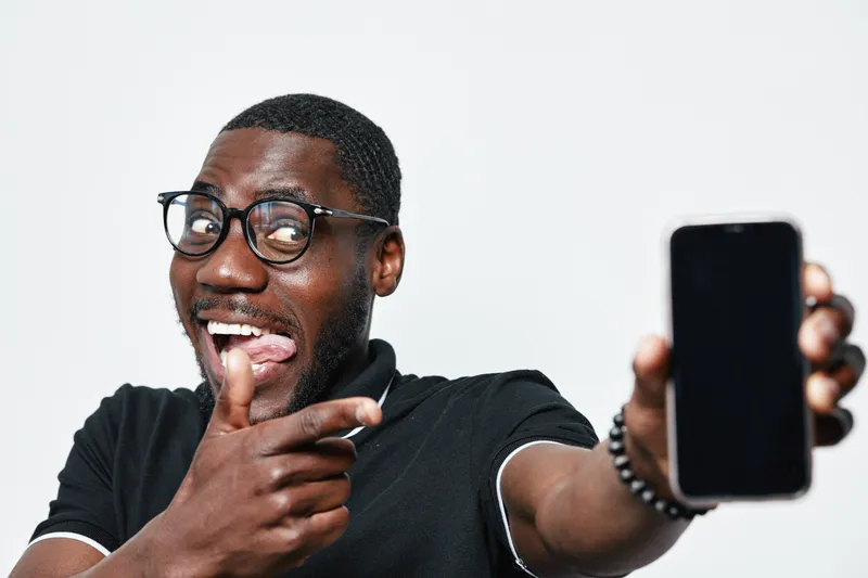 African American man with dark skin and glasses smiling and pointing at smartphone with blank screen in hand on white background. Casual clothing and happy expression with tech device.