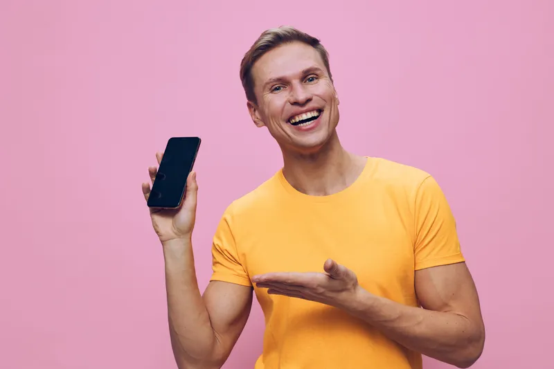 Smiling man with light skin and short brown hair wearing casual yellow t-shirt, holding smartphone in hand and gesturing with free hand, happy expression, studio portrait on pink background, lifestyle
