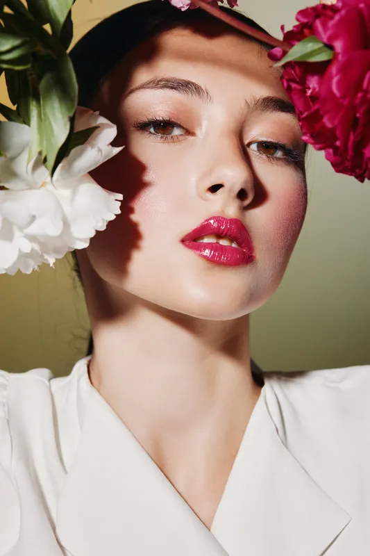 Close-up portrait of young woman with glossy pink lips and subtle eye makeup surrounded by vibrant flowers. Natural light highlights flawless skin and beauty concept.