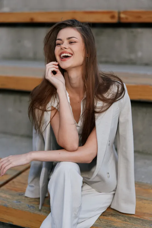 Happy woman in light business suit laughing outdoors with casual elegance, natural makeup, and relaxed pose on wooden stairs in soft neutral tones