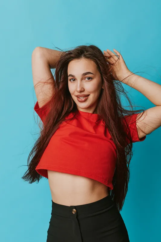 Young woman posing with hands in hair wearing red cropped top and black pants on blue background. Smiling female model with long hair and casual outfit in studio portrait.