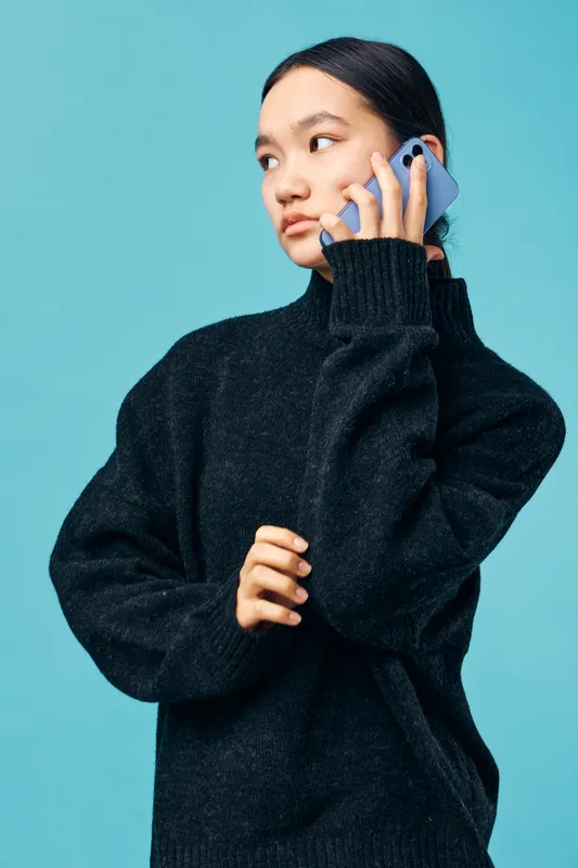 Young woman in black sweater holding smartphone to ear, looking sideways against blue background. Female model with casual outfit, communication and modern technology concept.