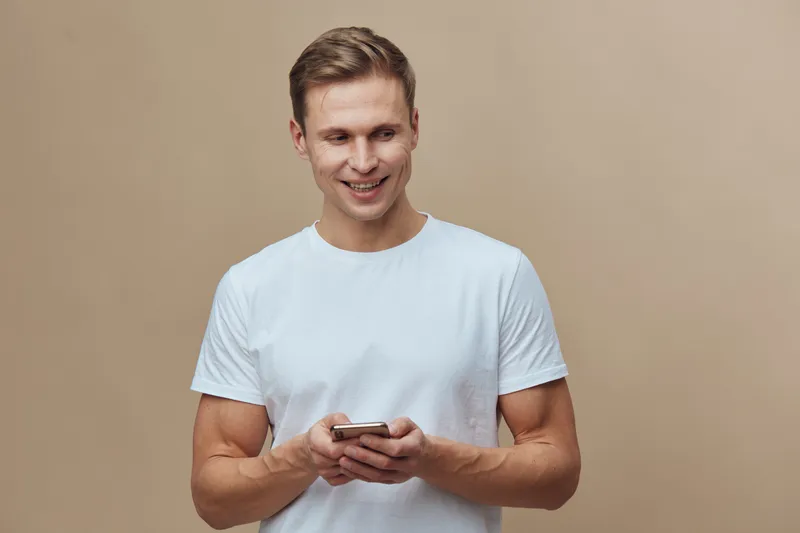 smiling man holding smartphone wearing white t-shirt looking aside on beige background digital nomad lifestyle communication concept
