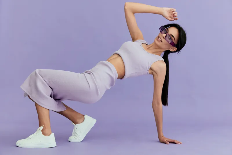 Young woman in lilac knitwear posing dynamically against lilac background, wearing white sneakers and purple sunglasses, showing modern style and confidence.