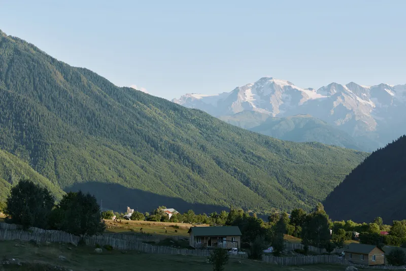 A tranquil mountain valley scene unfolds with green hills, distant snow capped peaks, and a clear blue sky. Meadows dot the landscape as rustic cottages sit beside stone walls and gentle sunlight