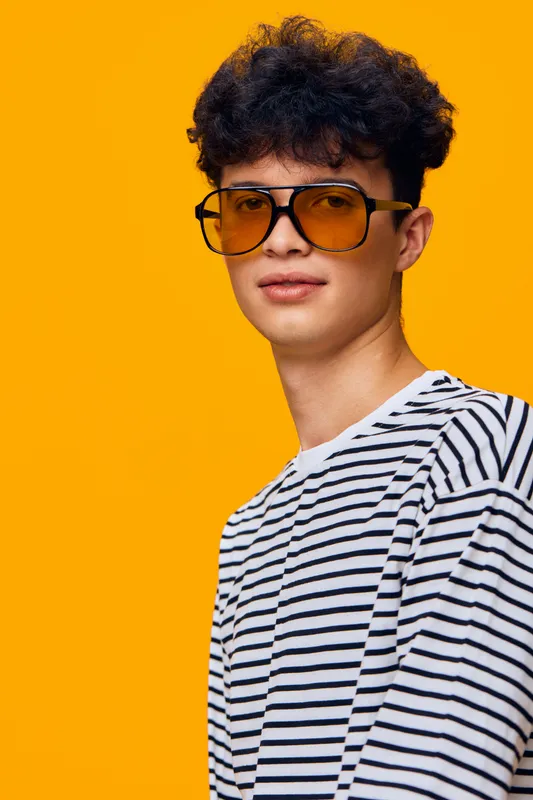 Young man wearing oversized yellow sunglasses and a striped shirt smiling confidently against a vibrant orange background in a stylish casual portrait.