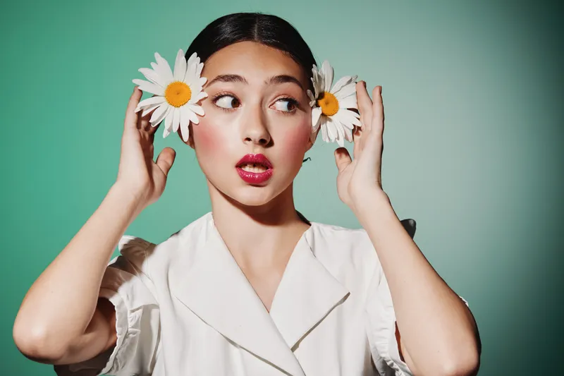 Young woman with smooth skin holds telephone receiver adorned with white daisies near ears. Creative fashion beauty concept with playful expression on green background.