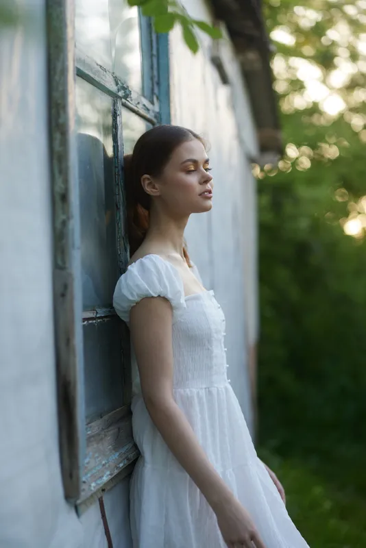 Young woman in a white dress standing by a rustic window in nature, exuding tranquility and grace Soft lighting enhances the serene atmosphere