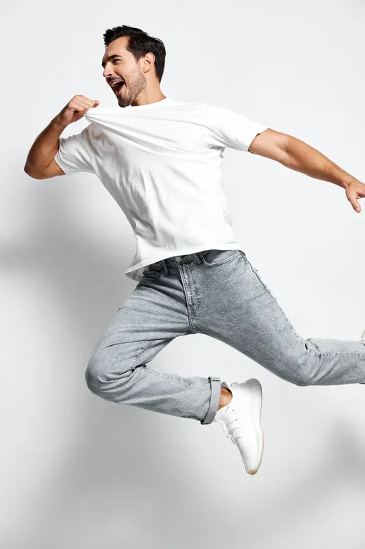 Happy energetic young man jumping in the air, wearing a casual white t-shirt and gray jeans, with a joyful expression on his face, against a minimalistic background.