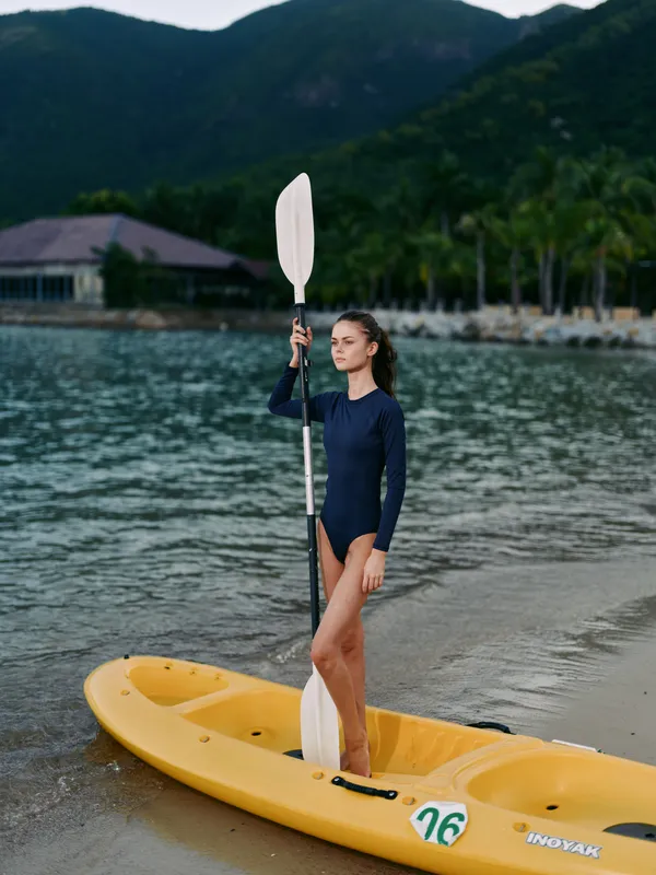 Young woman stands on a bright yellow kayak at the shoreline, holding a paddle upright in a navy swimsuit, with calm lake water and distant mountains.