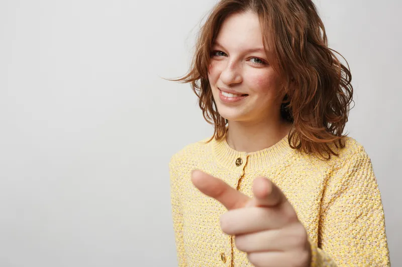 Smiling young woman with curly hair wearing yellow knitted sweater points finger at camera. Friendly gesture and positive emotion on light neutral background. Concept of confidence and connection.