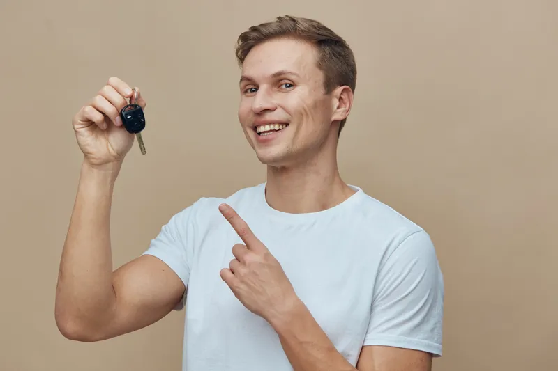 Smiling man with short brown hair holding car key and pointing at it, wearing casual white t-shirt, standing against plain beige background, studio shot, lifestyle concept, happiness, celebration, own