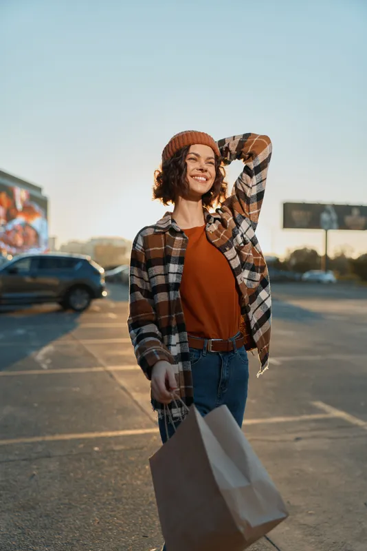 Woman shopping with smile in candid urban lifestyle scene, holding bag and wearing flannel and beanie at golden hour glow. Authenticity and mindful living with emotional storytelling vibe.