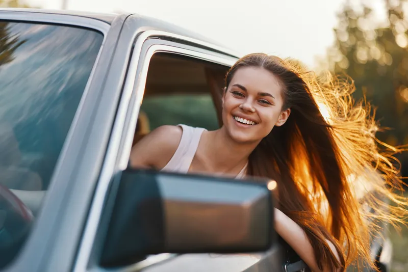Happy young woman with long hair enjoying a sunny day while leaning out of a car window, showcasing freedom and joy in the moment.