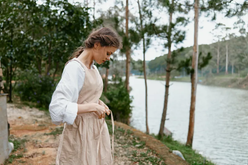 Young woman in a beige apron standing by a calm river, reflecting on nature with an introspective expression, ideal for healthy lifestyle concepts