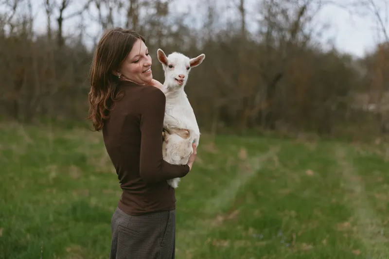 A young woman in a brown sweater holds a white baby goat in her arms while standing on a grassy path surrounded by trees on a cloudy day.