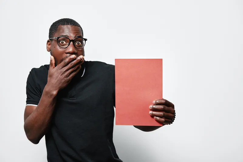 African American man with dark skin and glasses holding red paper with surprised expression on face in studio. Casual clothing and isolated white background for concept or message.