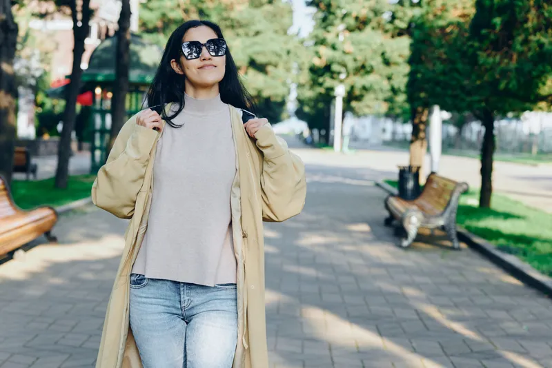 Woman in beige coat and denim walks through a sunlit park, stylish sunglasses and casual attitude convey confidence, fashion and outdoor elegance in a bustling urban setting with trees