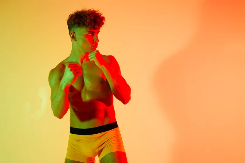 Athlete young man shirtless in active pose practicing boxing with intensity, expression and muscle definition under colorful lighting and studio composition for sport fitness.