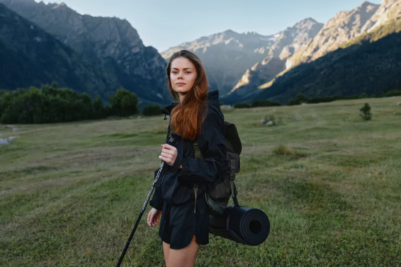 A young woman hiker stands in a wide alpine meadow with towering mountains in the background. She carries a backpack and trekking pole, dressed in outdoor gear, looking ahead with confidence and