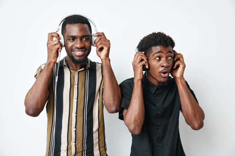 Two black men with dark skin wearing headphones enjoying music together, one smiling and the other surprised, standing against white background in casual shirts.
