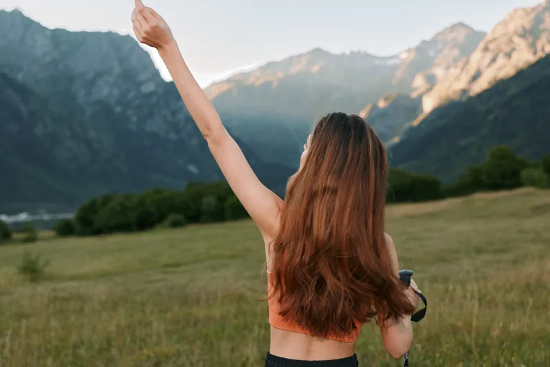 A young woman stands in a wide meadow with distant mountains, her arm raised in a celebratory gesture as sunlight sweeps across the tranquil landscape.