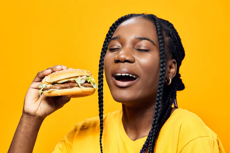 Happy young Black woman enjoying a delicious burger against a vibrant yellow background, representing a joyful food moment, taste satisfaction, and casual dining