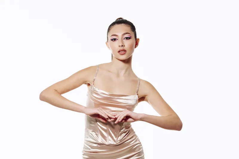 Stylish young woman poses confidently wearing silky slip dress. Her hair is tied back and makeup is creative with pink and purple stylish tones.