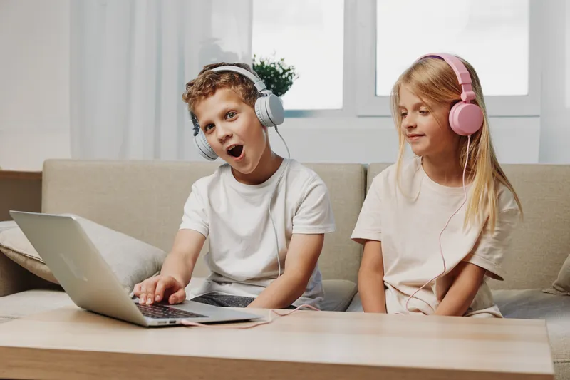 Two children wearing headphones sit on a sofa using a laptop, boy reacting excitedly and girl watching calmly in a bright living room. Concept of technology and childhood interaction.