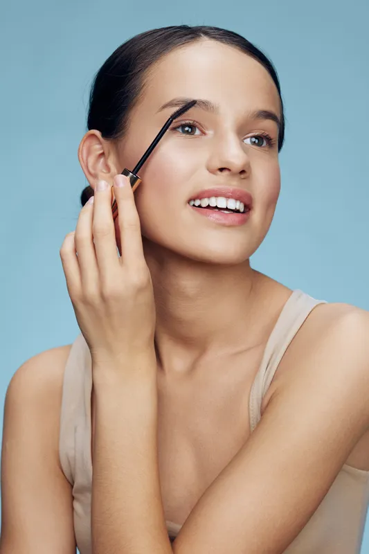 Smiling young woman applying brow gel, showcasing natural beauty and modern makeup trends against a soft pastel blue background, ideal for beauty and cosmetics themes