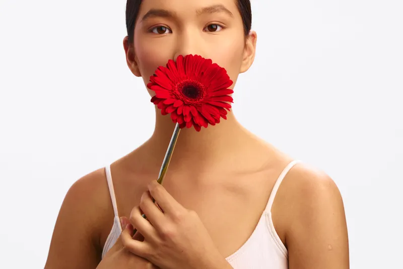 Asian woman holds a red flower near her lips, creating a calm, artistic portrait with soft studio lighting and a minimal background that enhances her serene presence.