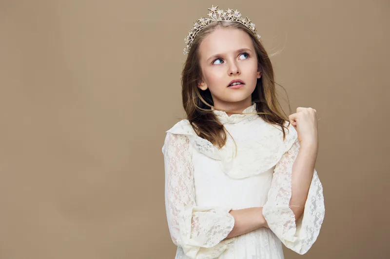 A young girl with a crown, wearing a white lace dress, gazes thoughtfully against a beige background, exuding innocence and elegance