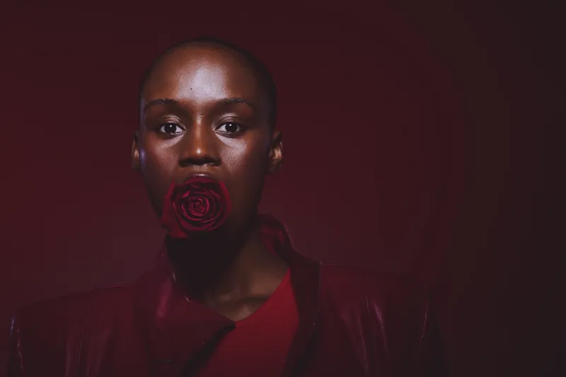 Closeup portrait of a dark skinned African woman with a shaved head holding a red rose in her mouth against a dark red background. Concept of beauty, elegance, and natural skin glow.