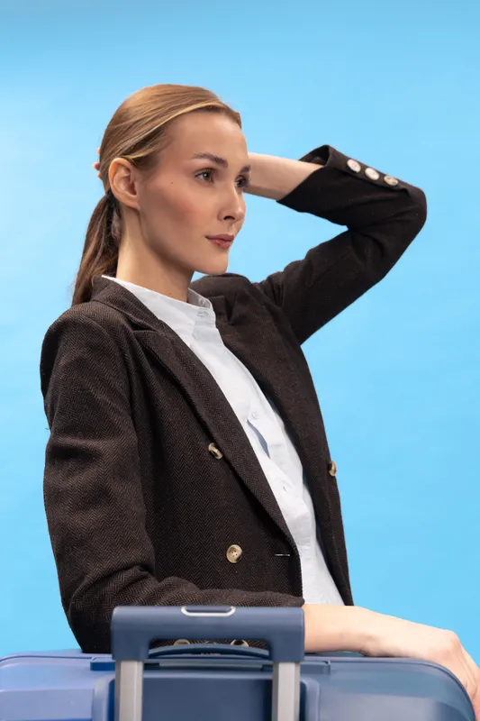 Young woman in dark blazer stands with hand on head looking sideways. Light blue background and travel suitcase visible. Concept of travel preparation, business trip, and thoughtful mood.