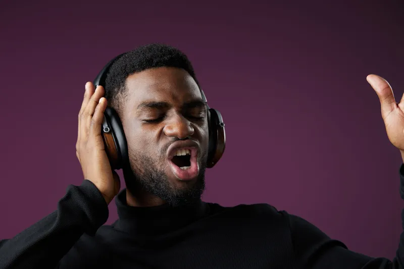 Young African man enjoying music with headphones, expressing joy and passion in a dark setting with purple background