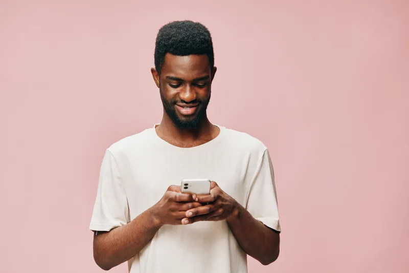 Young Black man smiling while using a smartphone, wearing a casual beige t shirt against a pink background, expressing happiness and engagement with technology