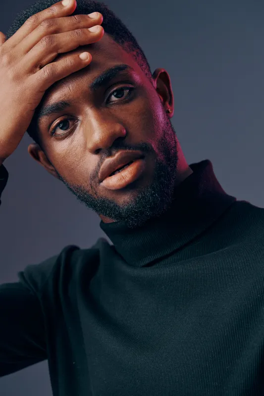 Young Black man with a thoughtful expression in a black turtleneck, highlighting introspection and emotion against a dark gray background