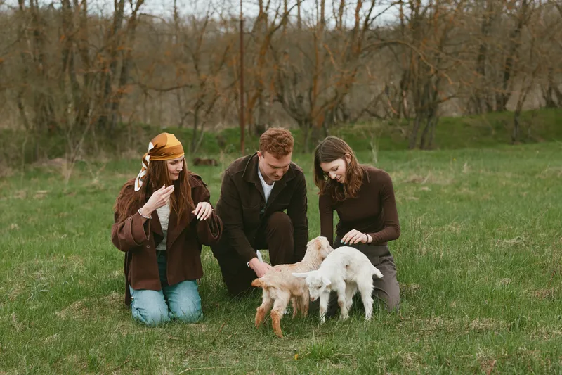 Three young adults enjoy a spring day outdoors playing with two baby goats on green grass in casual warm clothing creating a relaxed and natural lifestyle moment.