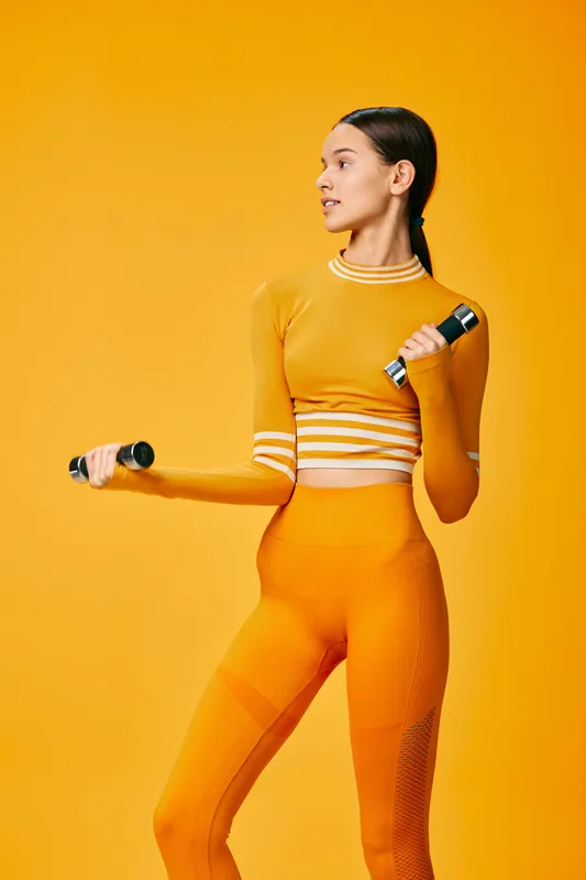 Vibrant portrait of a woman in orange athletic wear holding dumbbells, showcasing fitness, strength and balance in a bright studio setting with bold color.
