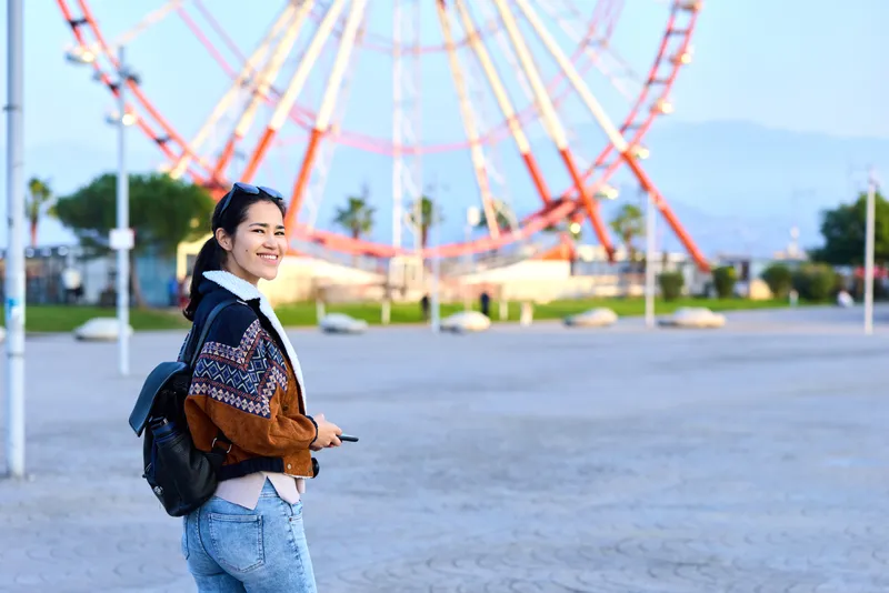 Woman with backpack at an amusement park near a ferris wheel, smiling while exploring outdoors on a sunny day, casual jacket and denim, capturing a candid travel moment in a bright scenic setting
