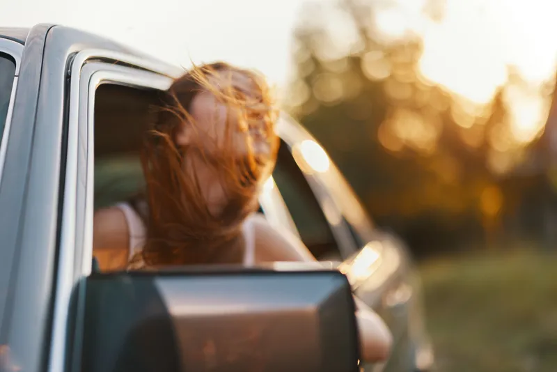 Young woman enjoying a road trip with wind in her hair, capturing the essence of freedom and adventure during sunset. Ideal for travel and lifestyle themes.