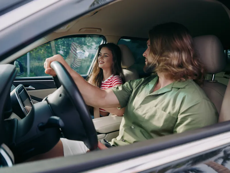 A joyful couple sits inside a modern car, smiling and sharing a moment as the driver handles the wheel, creating a warm, relaxed driving scene on a sunny day.