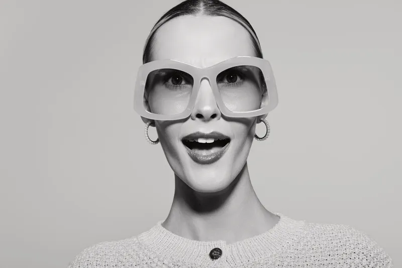 Young white woman with pale skin and slicked hair wearing large glasses smiles with open mouth in studio.